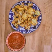 Overhead view of golden roasted cauliflower florets on blue and white patterned plate with spicy brava sauce in orange checkered bowl.