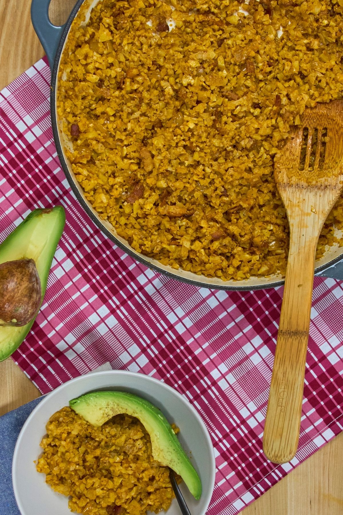 Close-up of golden cauliflower yellow rice with bacon pieces in braiser with avocado half and individual serving on white bowl.