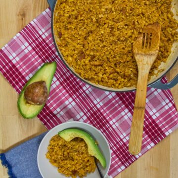 Overhead view of golden cauliflower yellow rice with bacon in braiser and white bowl with avocado slices on red plaid towel.