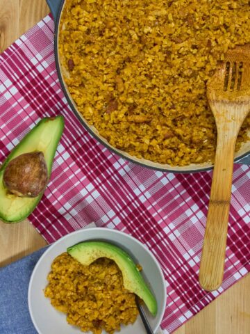 Overhead view of golden cauliflower yellow rice with bacon in braiser and white bowl with avocado slices on red plaid towel.