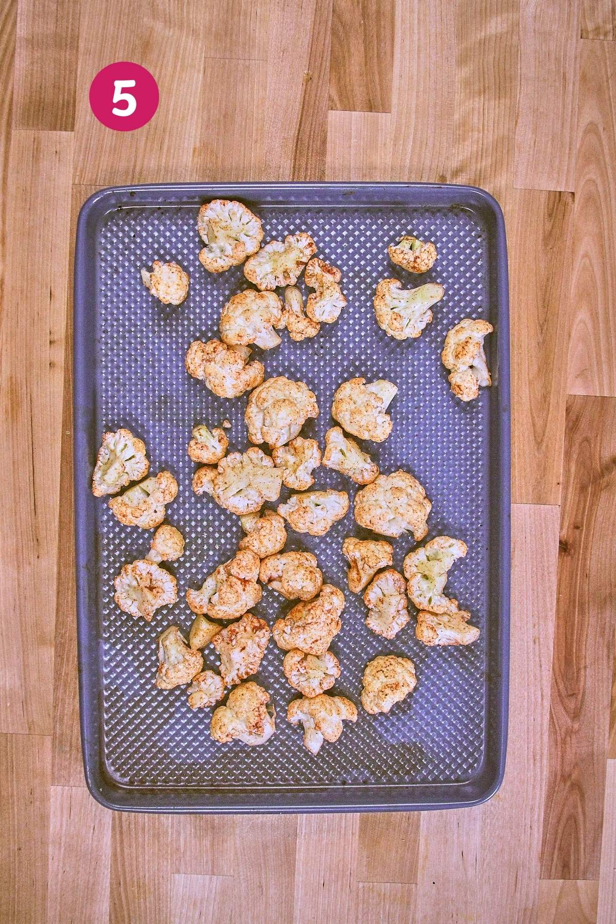 Overhead view of seasoned cauliflower florets arranged on dark perforated baking sheet ready for roasting, labeled with step 5.