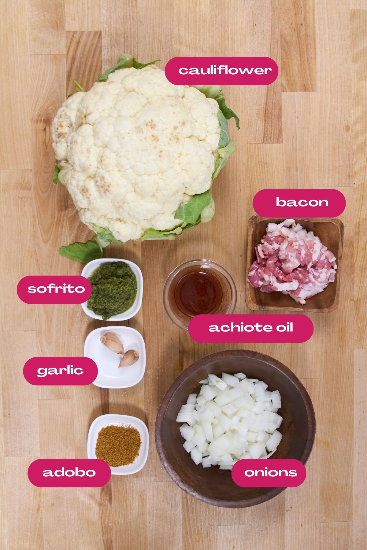 Overhead view of ingredients for cauliflower yellow rice including whole cauliflower head, diced bacon, sofrito, achiote oil, garlic cloves, diced onions and adobo seasoning in separate bowls.