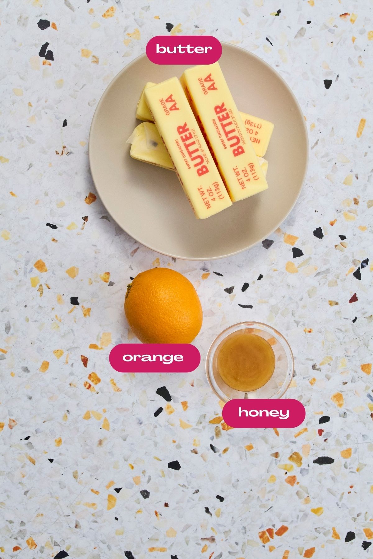 Orange honey butter ingredients: sticks of butter, fresh orange, and honey in a glass bowl with pink labels.