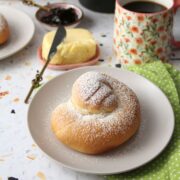 Pan de Mallorca roll dusted with powdered sugar on a gray plate, with butter, jam, and a mug of coffee in the background.
