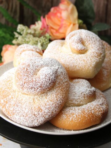 Pan de Mallorca rolls dusted with powdered sugar stacked on a plate on a cake stand, with flowers in the background.