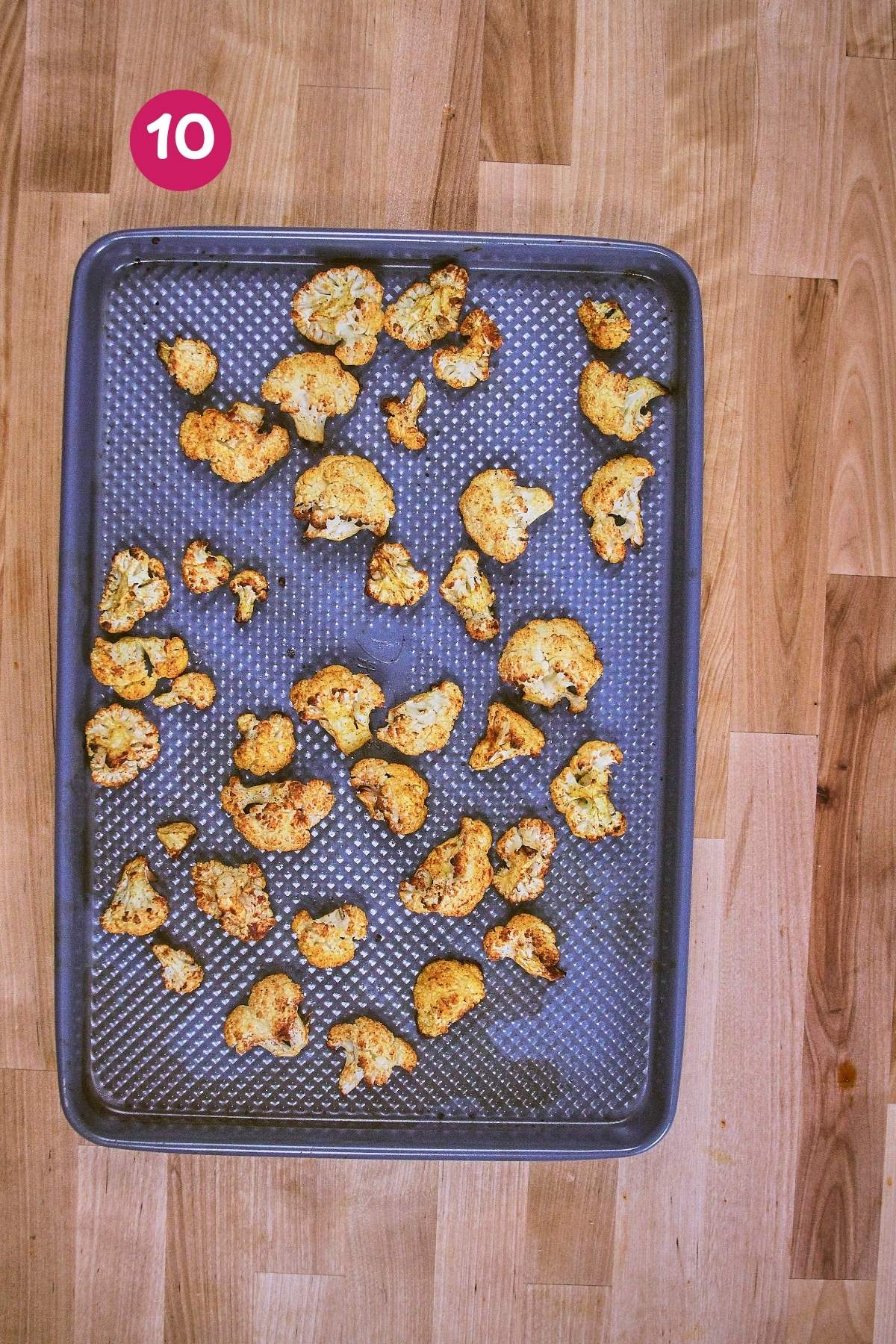 Overhead view of golden roasted cauliflower florets on dark perforated baking sheet after roasting, labeled with step 10.