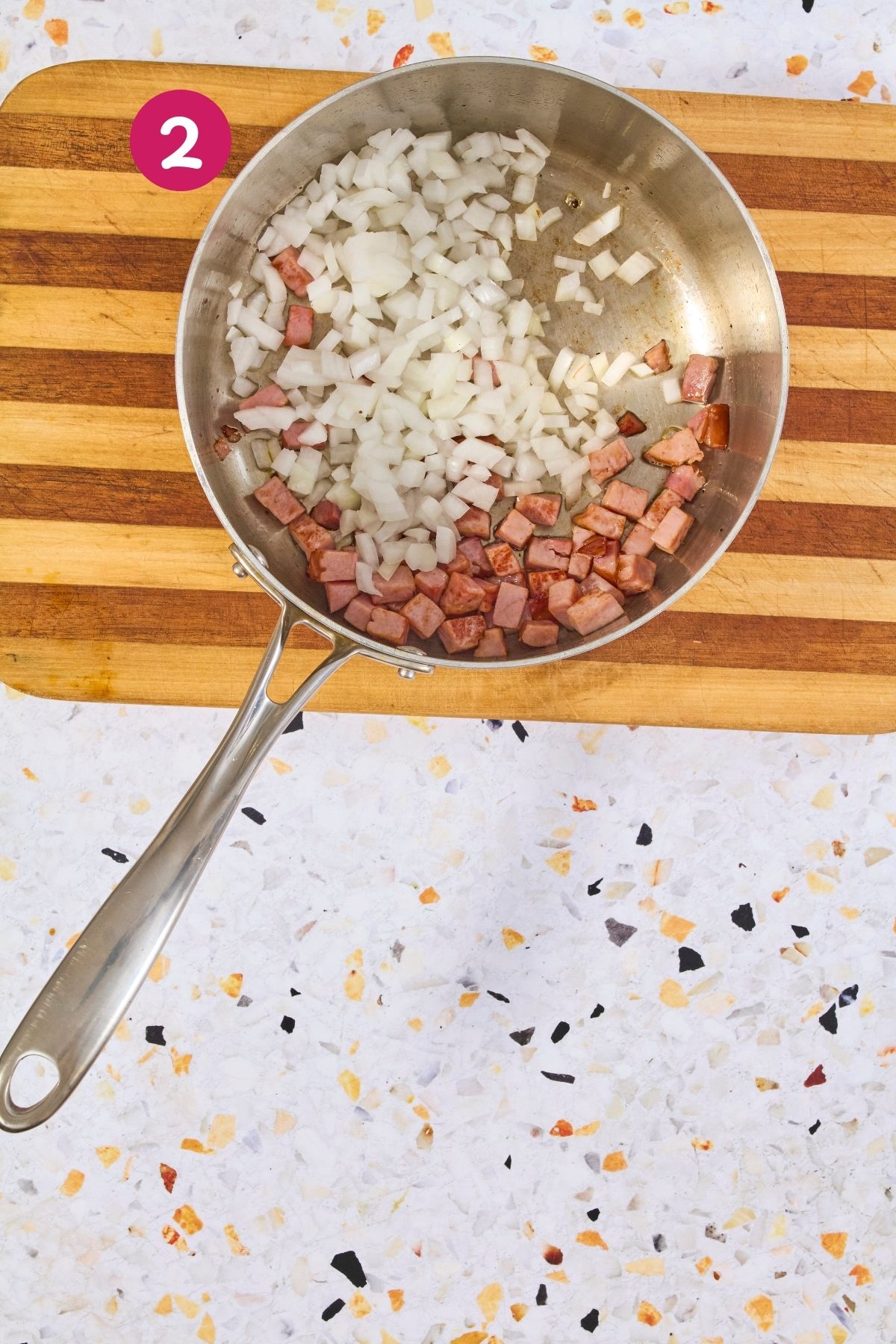 Overhead view of diced onions being sautéed with browned ham pieces in a stainless steel skillet, labeled with step 2.