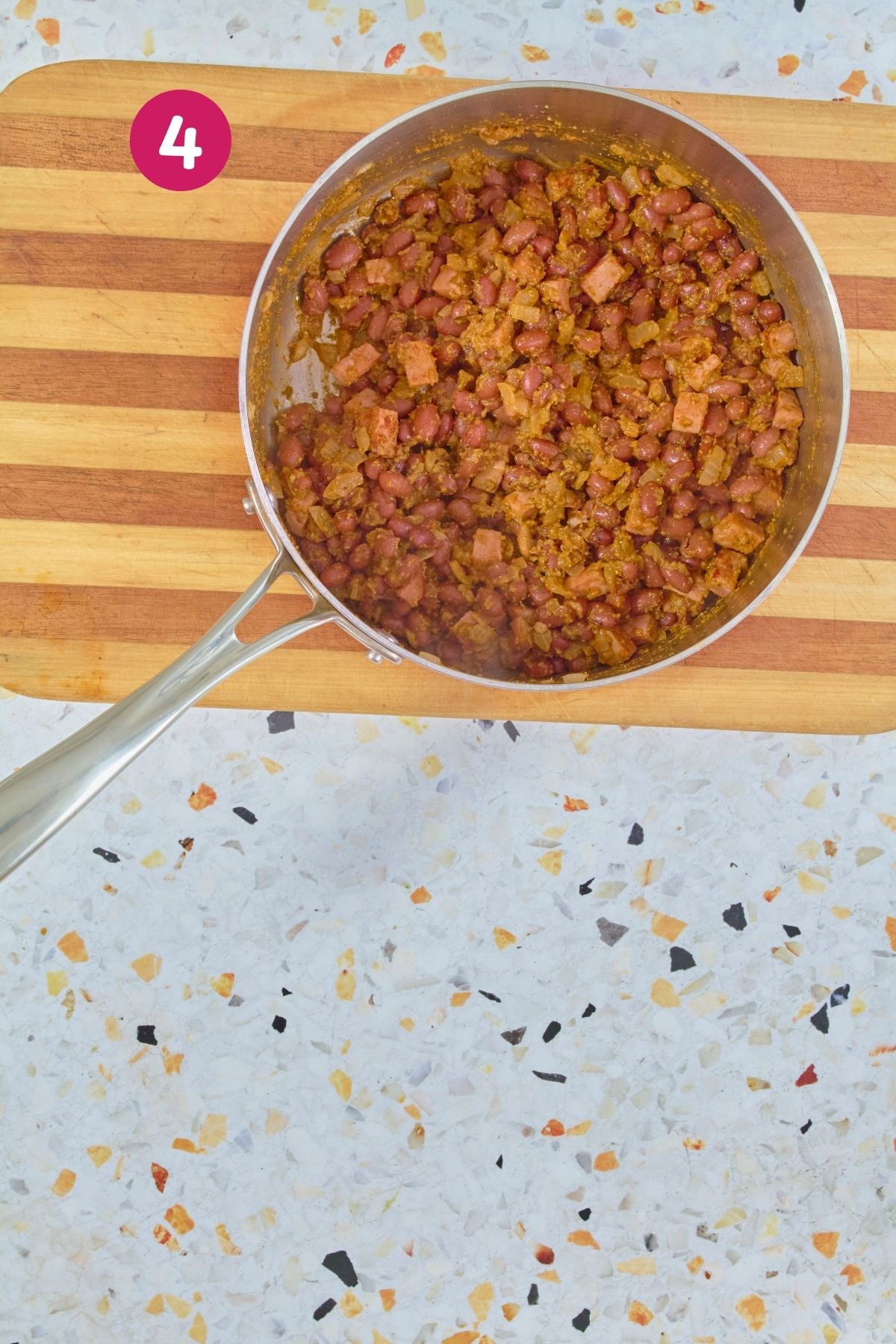 Overhead view of pink beans, ham, and onions seasoned with sofrito and garlic paste in a stainless steel skillet, labeled with step 4,