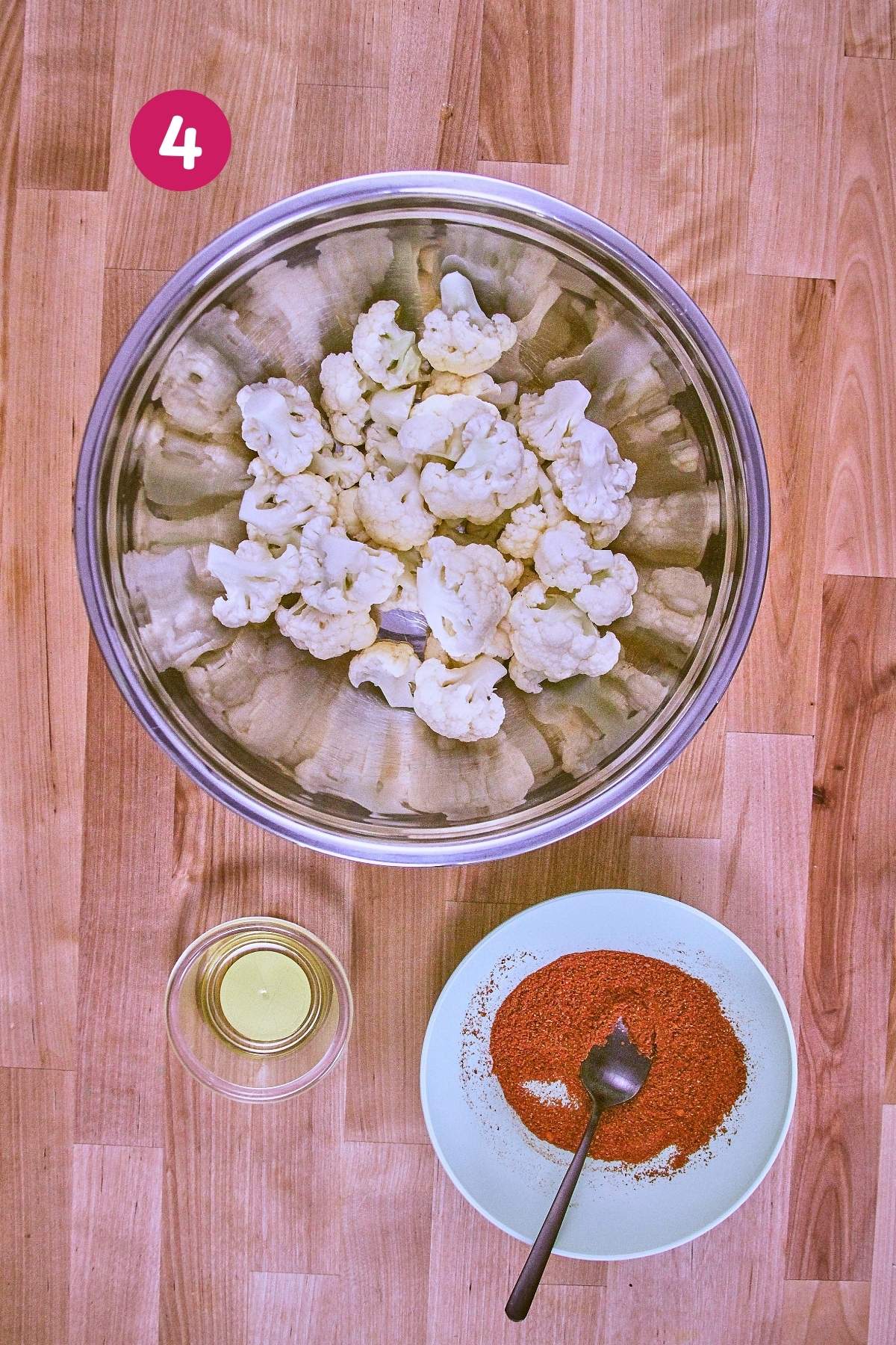 Overhead view of cauliflower florets in a bowl with spice mixture and olive oil beside it, ready to be tossed, labeled with step 4.