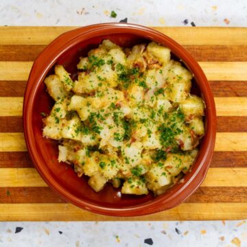 Yuca salad with crispy bacon, fresh herbs, and warm vinaigrette served in a rustic terracotta bowl on a wooden cutting board.