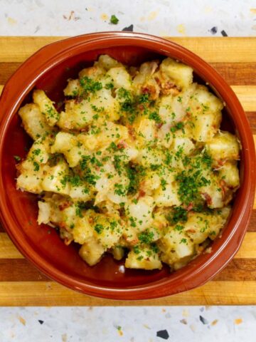 Yuca salad with crispy bacon, fresh herbs, and warm vinaigrette served in a rustic terracotta bowl on a wooden cutting board.