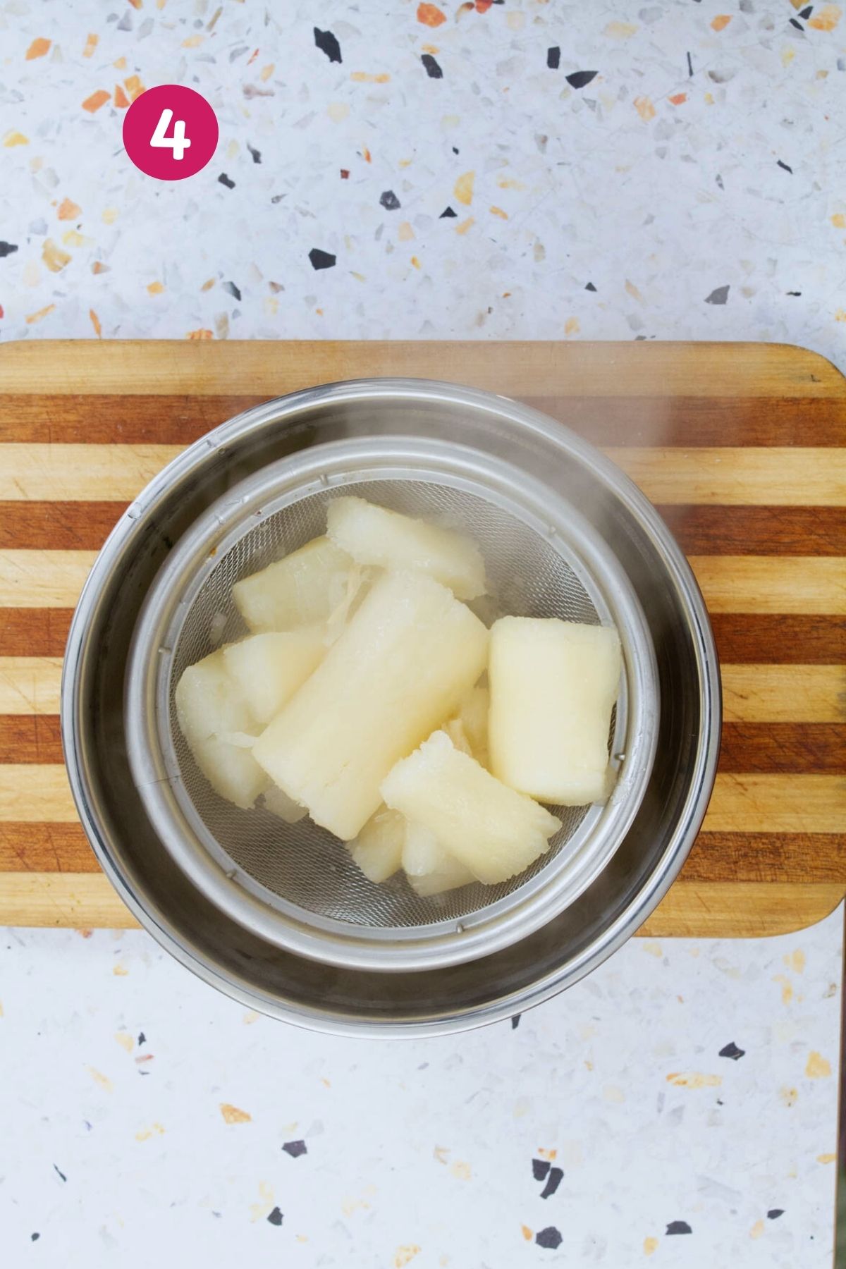 Chunks of peeled yuca root are boiling in a stainless steel strainer basket over a pot of water.