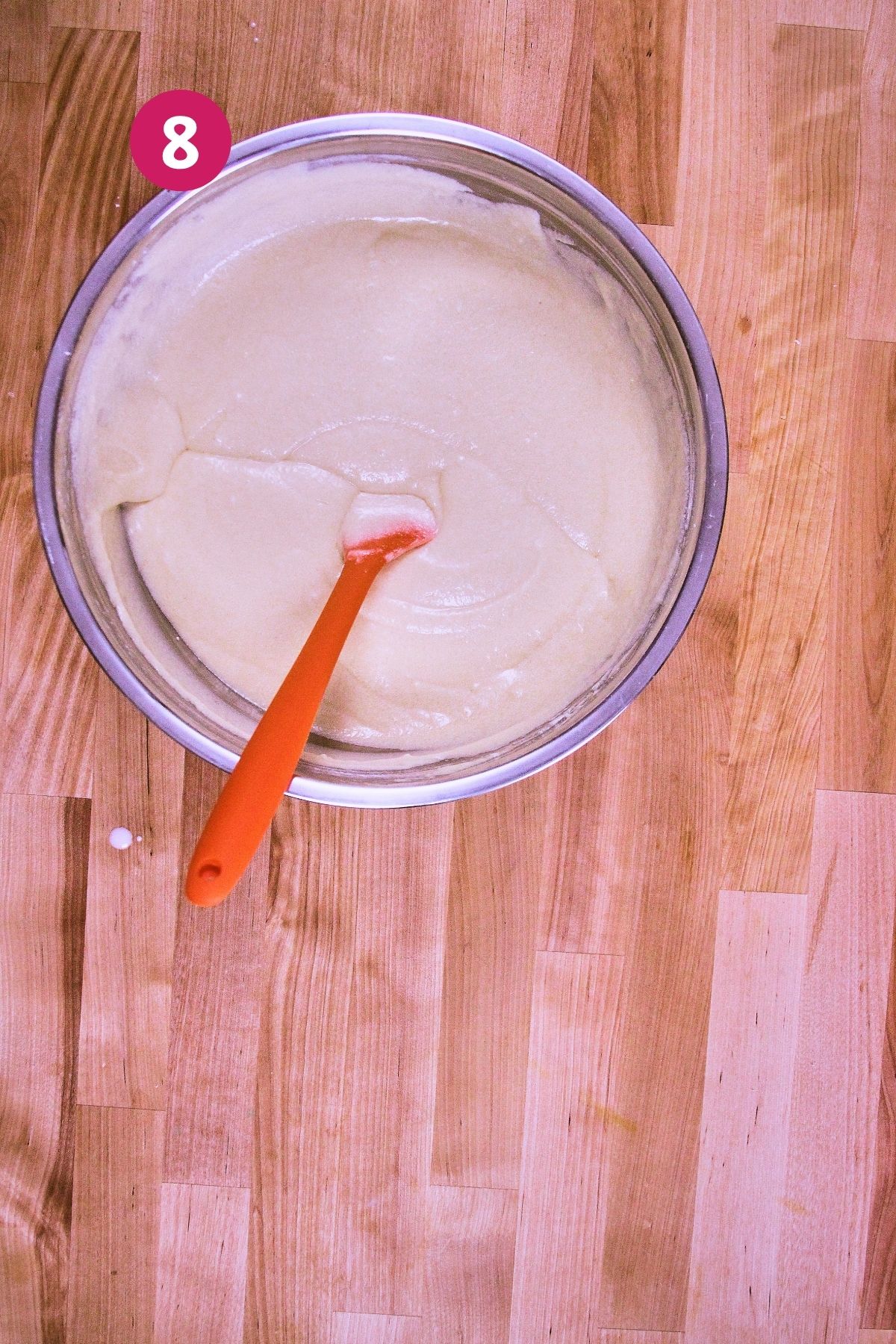 Finished vanilla cake batter in a metal mixing bowl with an orange silicone spatula, ready to be poured over the flan custard.