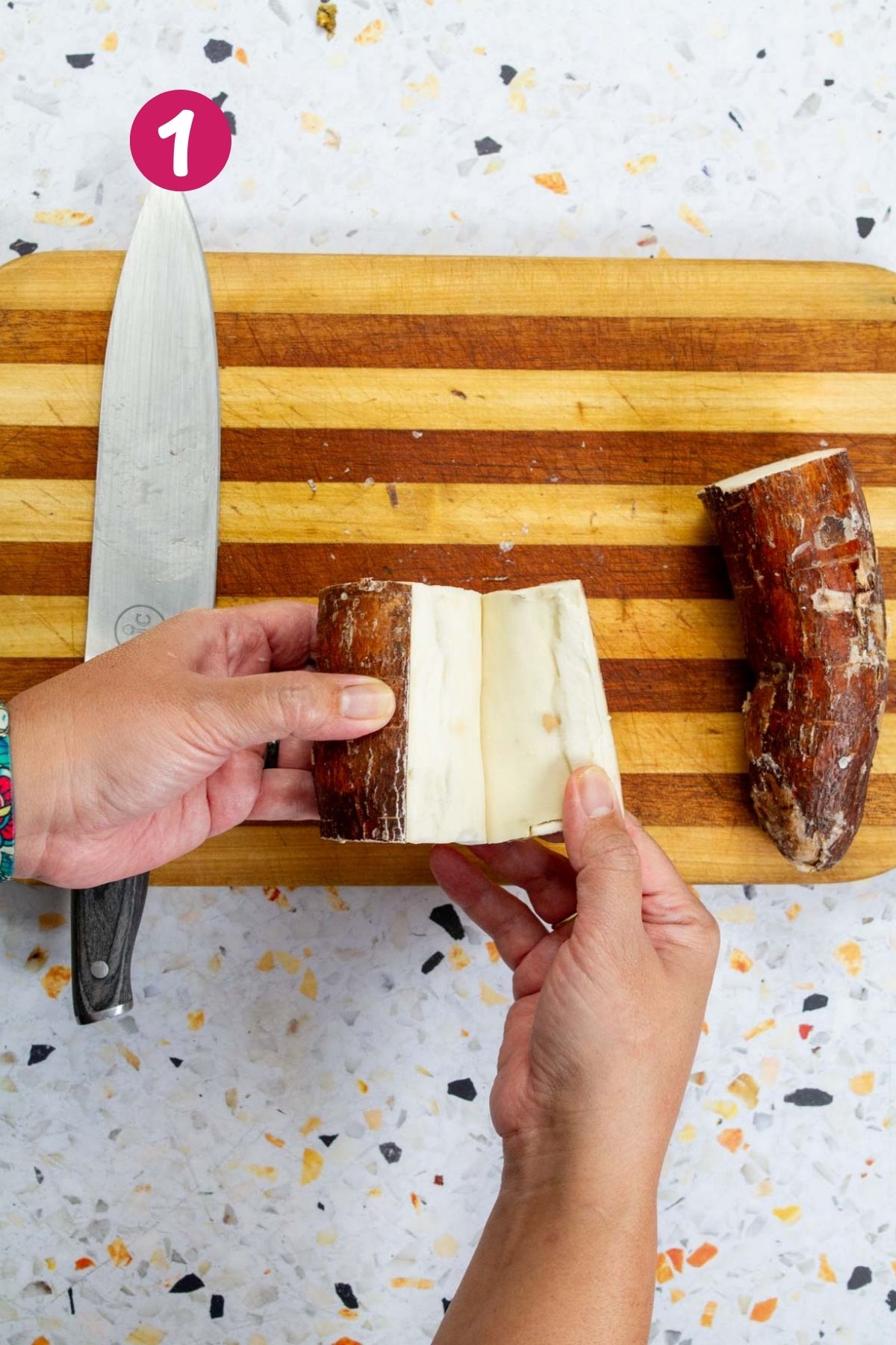 Hands peeling thick brown skin from fresh yuca root with a chef's knife on a striped wooden cutting board.