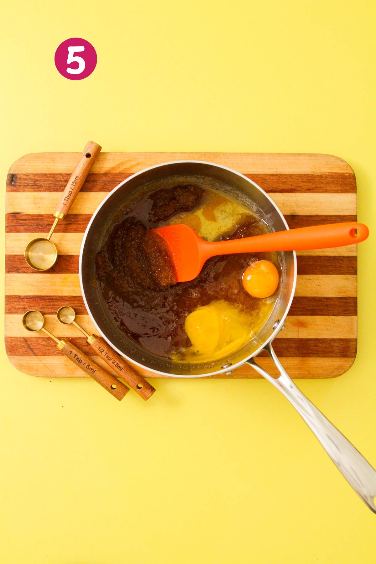 Egg and egg yolk added to brown butter and sugar mixture in a stainless saucepan, with measuring spoons on a striped wooden cutting board.