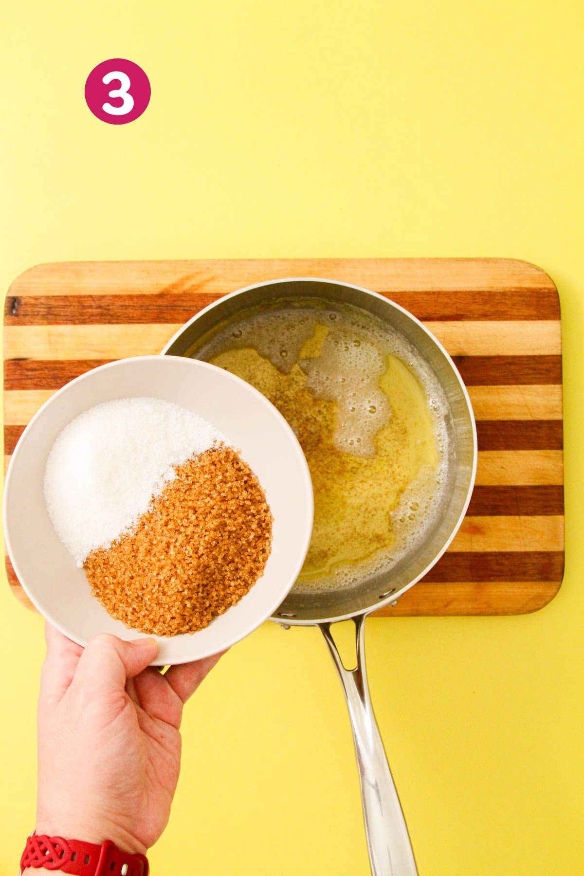 Hand holding a white bowl with granulated and brown sugar over a saucepan of browned butter on a striped wooden cutting board.