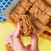 Hand holding a peanut butter chocolate chip blondie broken in half to reveal a dense, chewy interior with visible chocolate chips, pecans, and peanut butter pieces, with a full tray of cut blondies on parchment paper in the background.