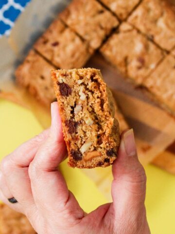 Hand holding a peanut butter chocolate chip blondie broken in half to reveal a dense, chewy interior with visible chocolate chips, pecans, and peanut butter pieces, with a full tray of cut blondies on parchment paper in the background.
