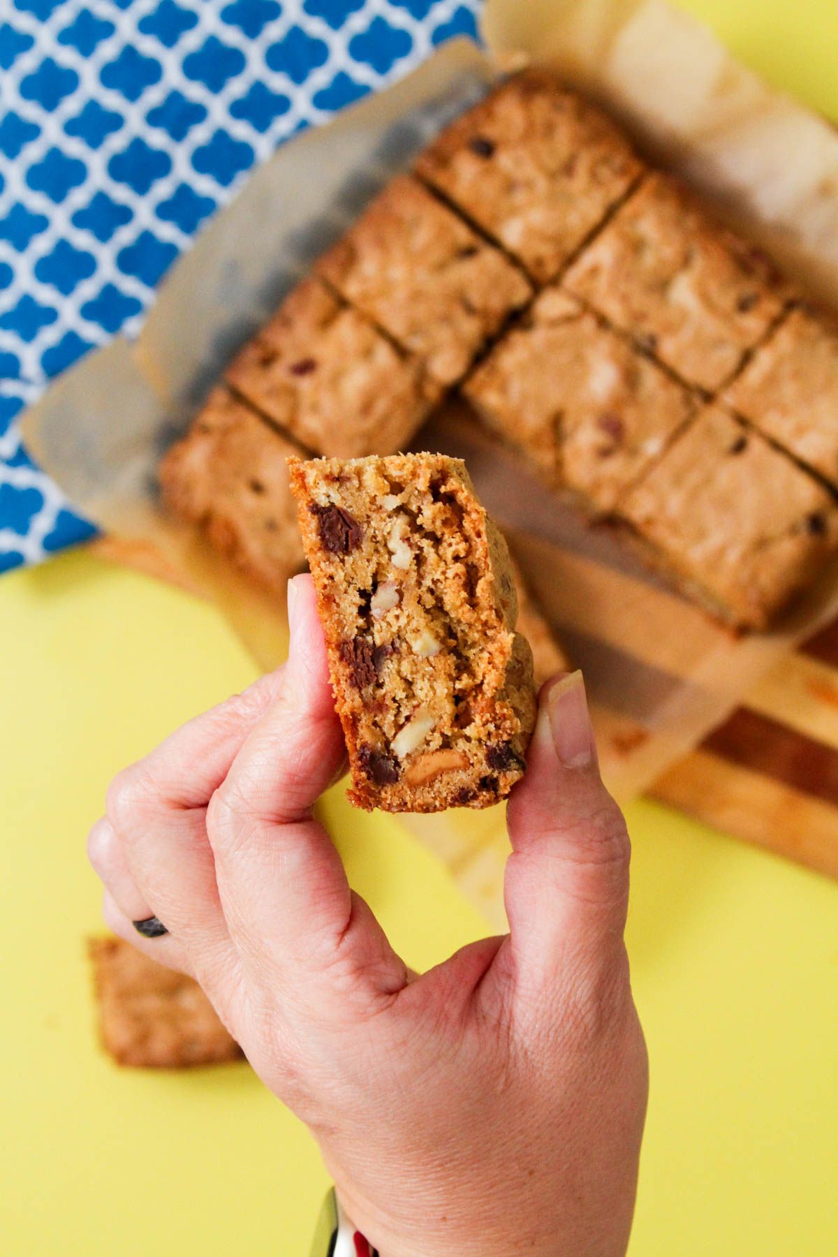 Hand holding a peanut butter blondie cut in half showing a dense, chewy interior studded with chocolate chips and pecans, with a full tray of cut blondies on parchment paper in the background.