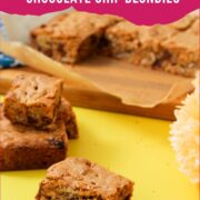 Peanut butter chocolate chip blondies cut into squares on a yellow surface with parchment paper and a yellow flower, with a pink and white Pinterest graphic overlay reading "Chewy and Buttery Classic Peanut Butter Chocolate Chip Blondies."