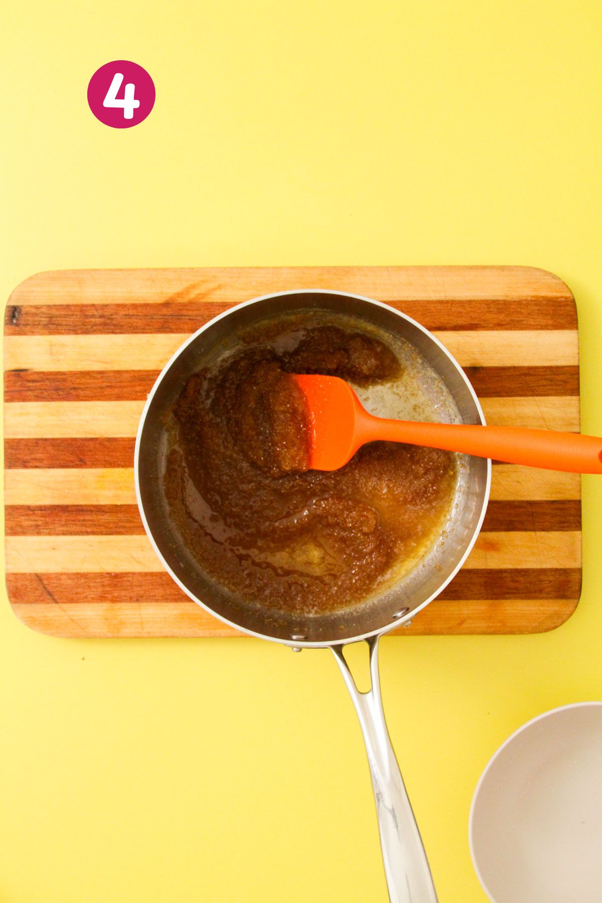Brown butter and sugar mixture cooling in a stainless saucepan with an orange rubber spatula, on a striped wooden cutting board.