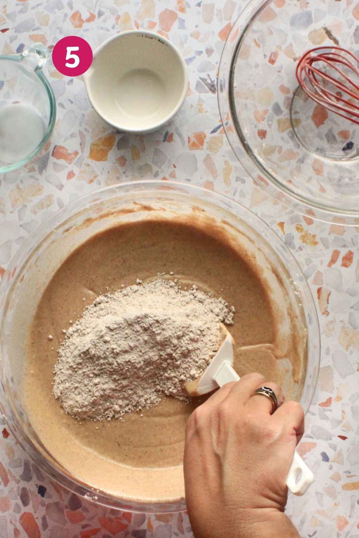 Hand folding the final addition of spiced flour into hojaldre añasqueño batter with a spatula.