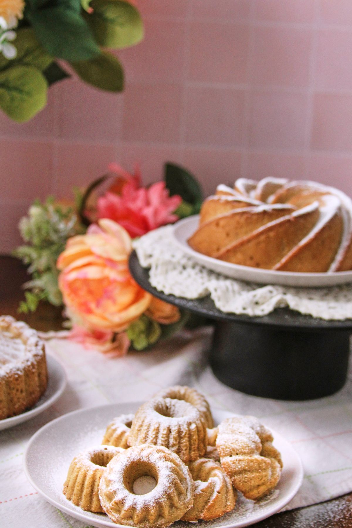 Puerto Rican spice cake hojaldre añasqueño displayed as mini bundts on a white plate with a spiral bundt on a cake stand, surrounded by pink and orange flowers.