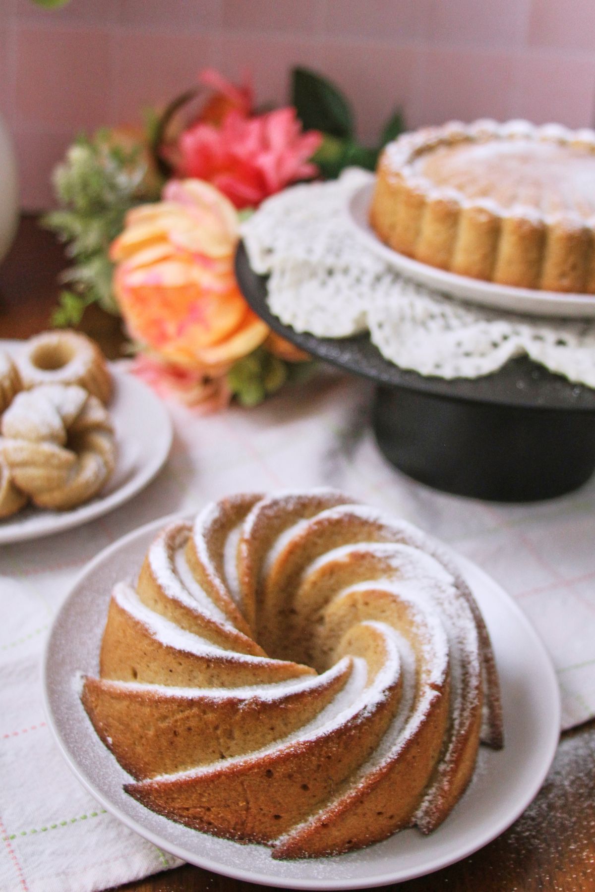 Hojaldre añasqueño Puerto Rican spice cake spiral bundt dusted with powdered sugar, with mini bundts and a Charlotte-style cake in the background.