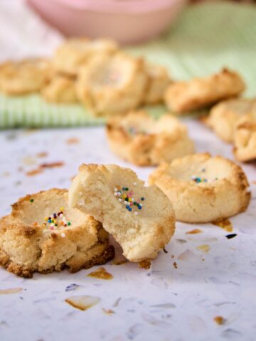 Close-up of low-carb polvorones broken open on a terrazzo surface showing their tender crumb, with more cookies and a pink bowl in the background.