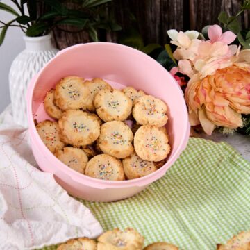 Low-carb polvorones with rainbow sprinkles piled in a pink bowl surrounded by peach flowers, green gingham cloth, and white linen on a styled surface.
