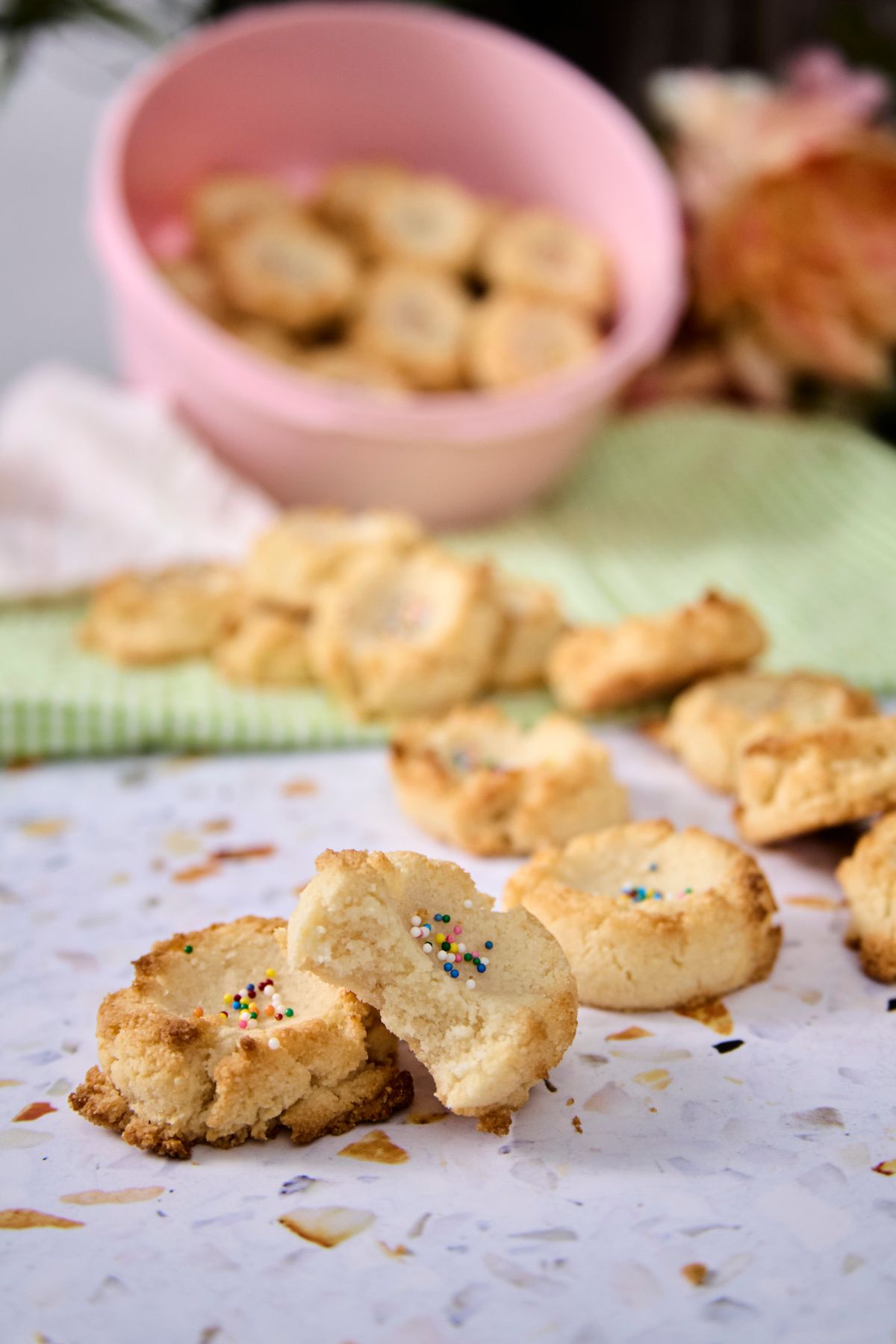 A low-carb polvorón broken in half, showing its tender interior with rainbow sprinkles, surrounded by more cookies on a terrazzo surface with a pink bowl in the background.