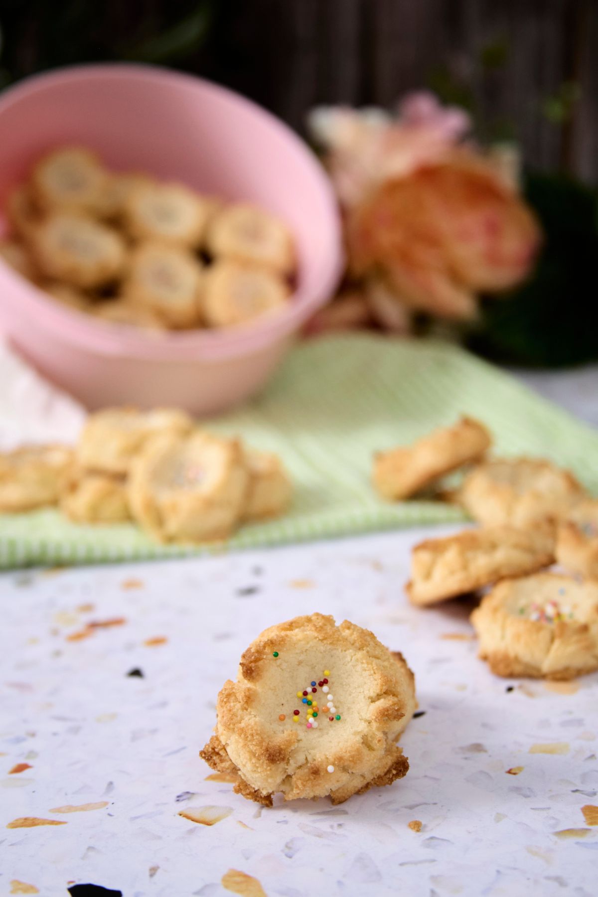 A single low-carb polvorón with rainbow sprinkles on a terrazzo surface with more cookies and a pink bowl of polvorones in the background.