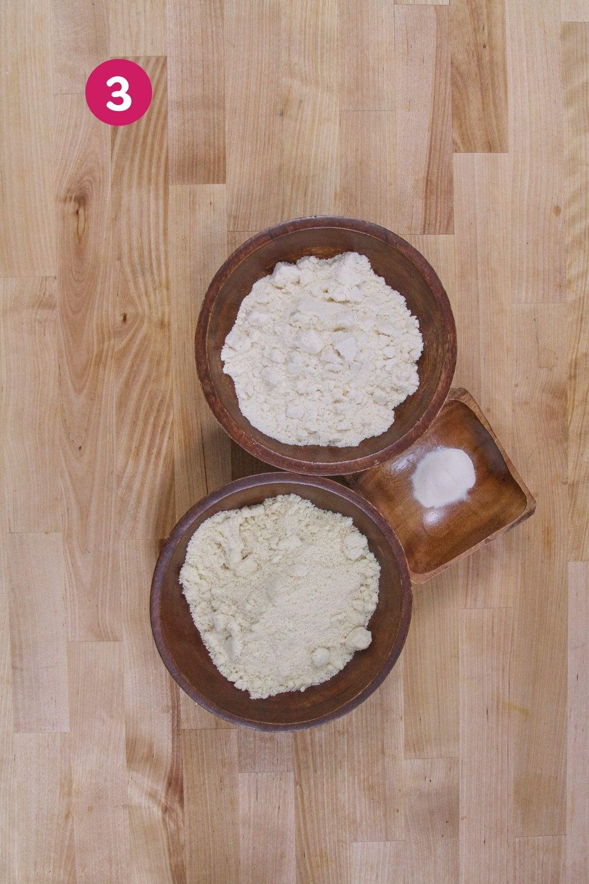 Two wooden bowls of coconut flour and almond flour with a small bowl of xanthan gum on a wooden surface.