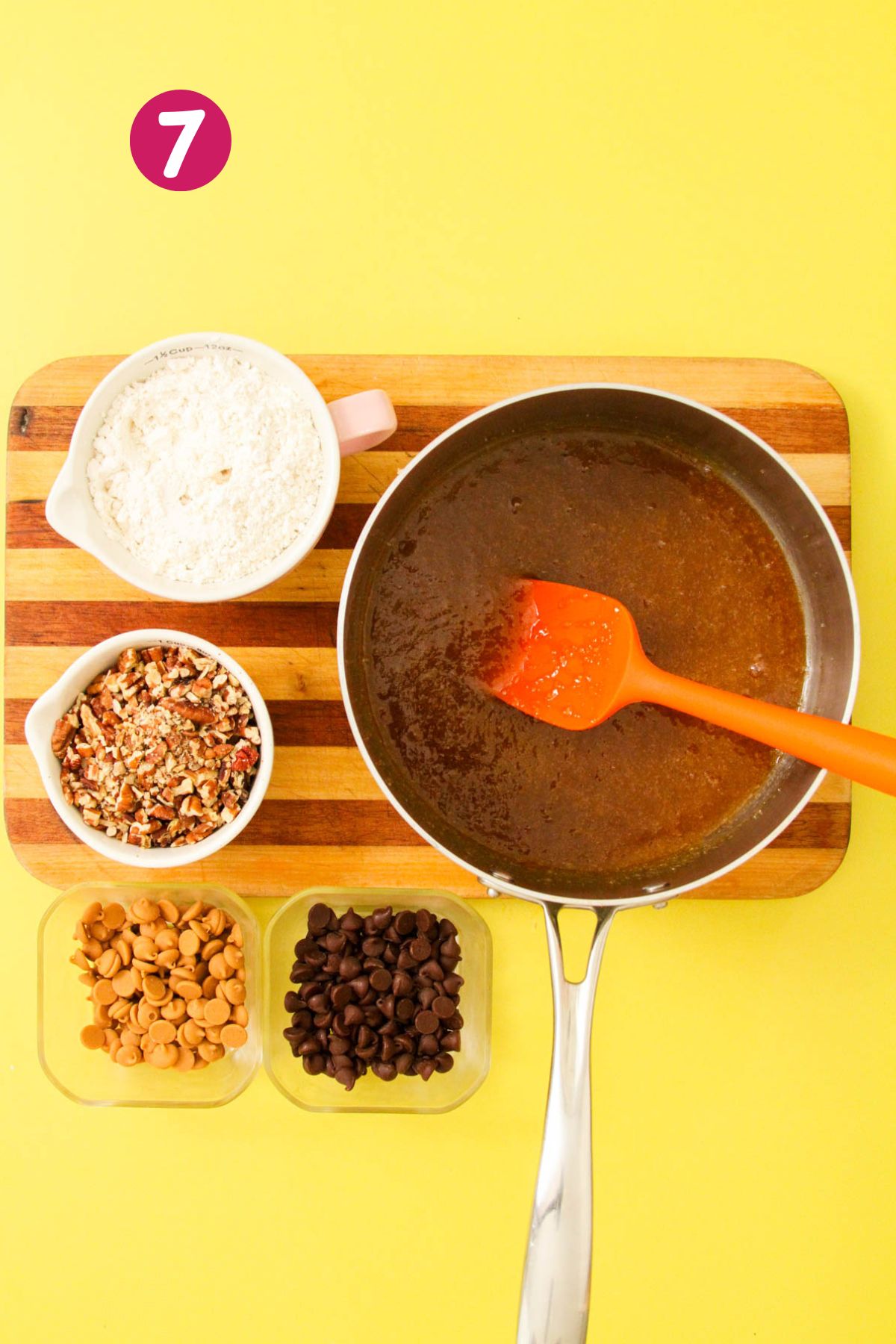 Wet blondie batter in a stainless saucepan surrounded by bowls of flour, chopped pecans, peanut butter chips, and chocolate chips on a striped wooden cutting board.