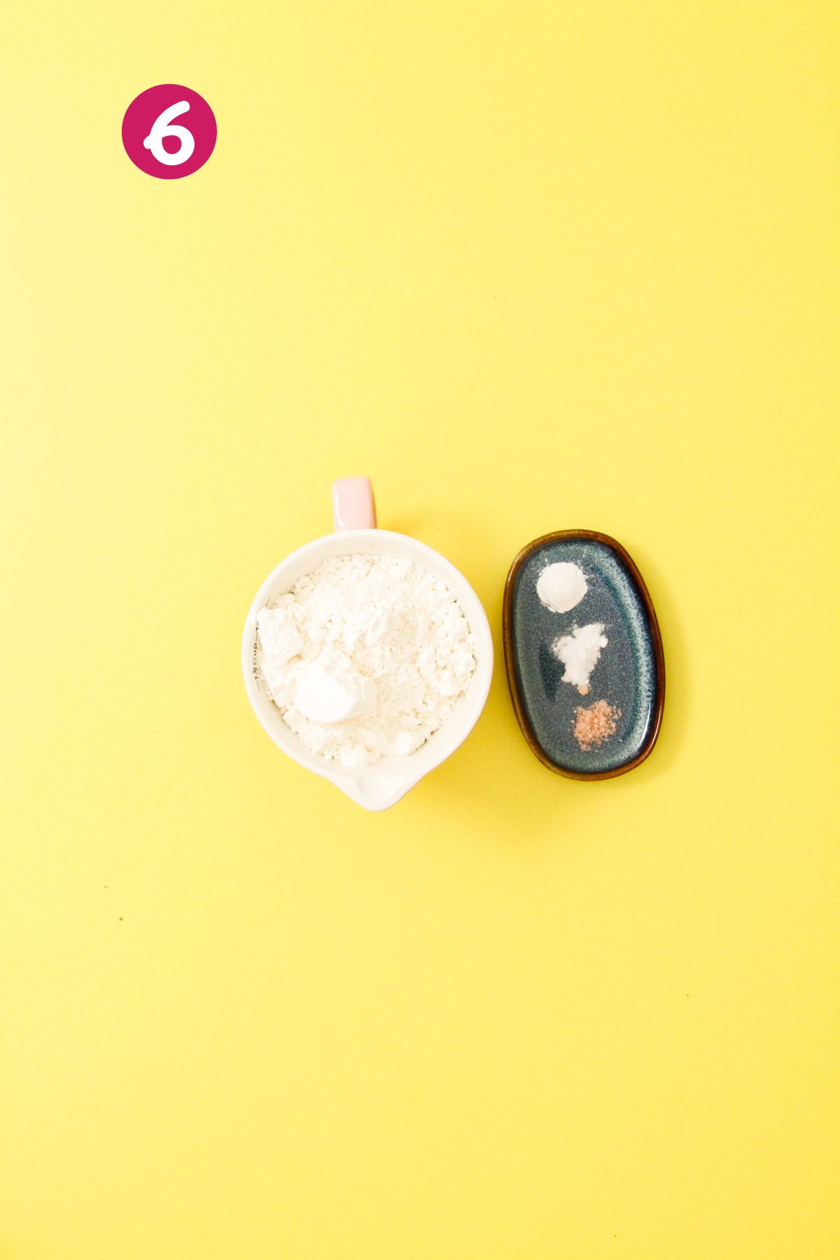 Small white measuring cup filled with all-purpose flour mixed with leavening agents beside a dark ceramic dish holding salt, baking soda, and baking powder on a yellow background.