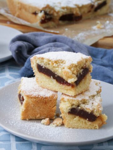 Three stacked panetela de guayaba bars on a gray plate showing the guava paste layer, with a blue linen and striped wood cutting board in the background.