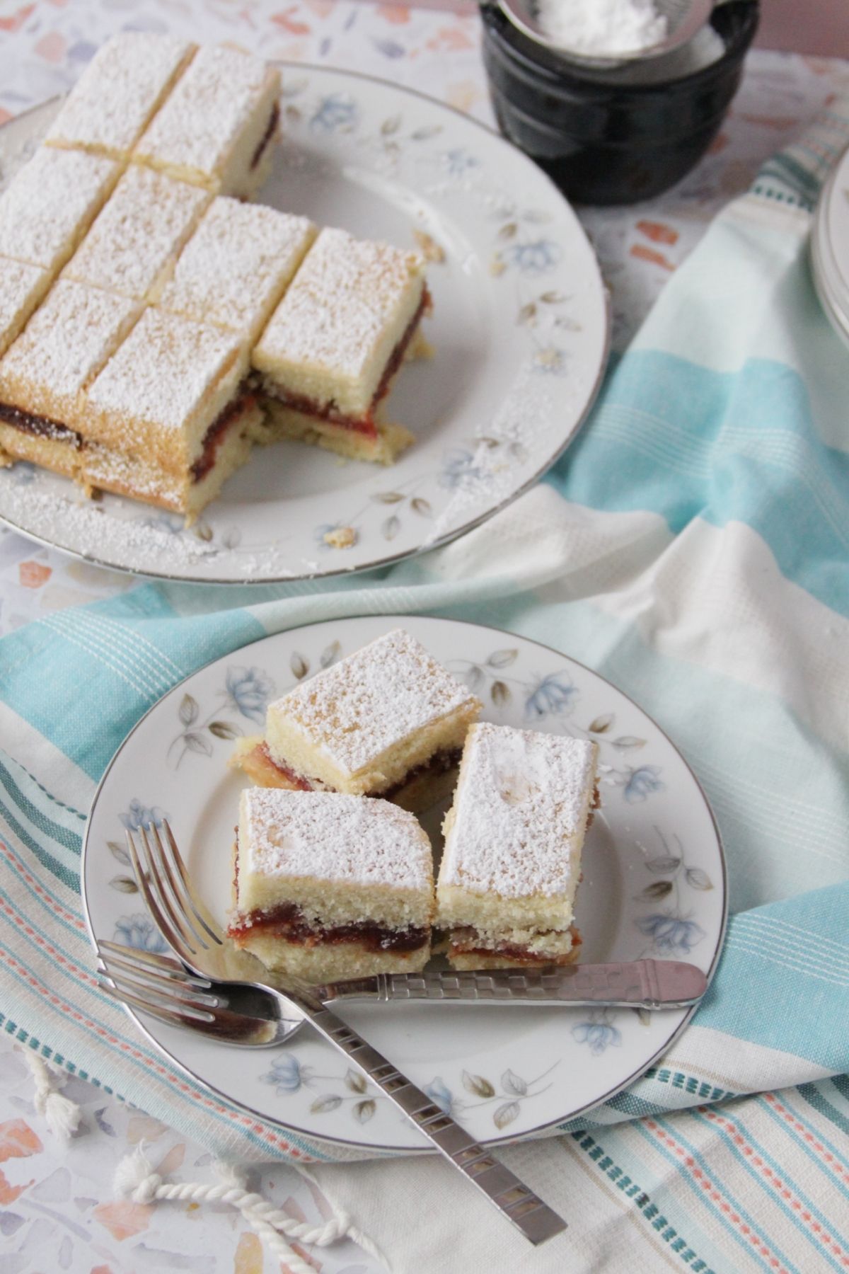 Two plates of panetela de guayaba bars dusted with powdered sugar, showing the guava paste layer, served on floral china with a fork and knife on a blue striped linen.
