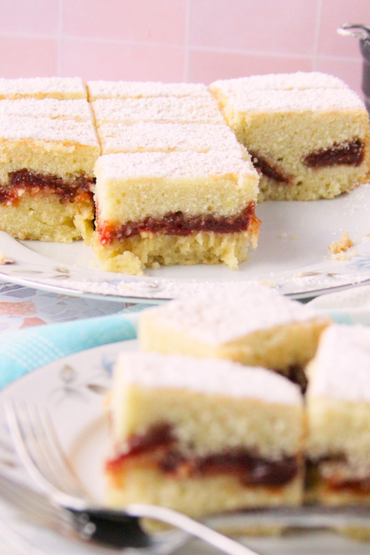 Close-up of panetela de guayaba bars on floral china showing the soft crumb and guava paste layer, with additional bars on a second plate in the background against a pink tile wall.