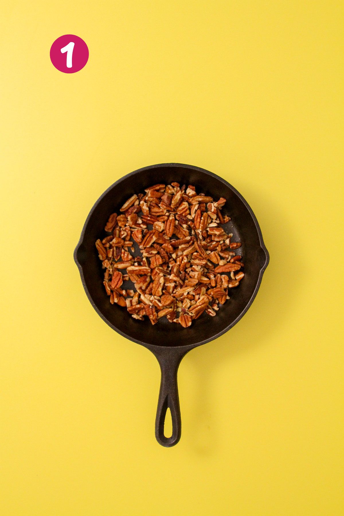 Chopped pecans toasting in a small cast iron skillet on a yellow background.