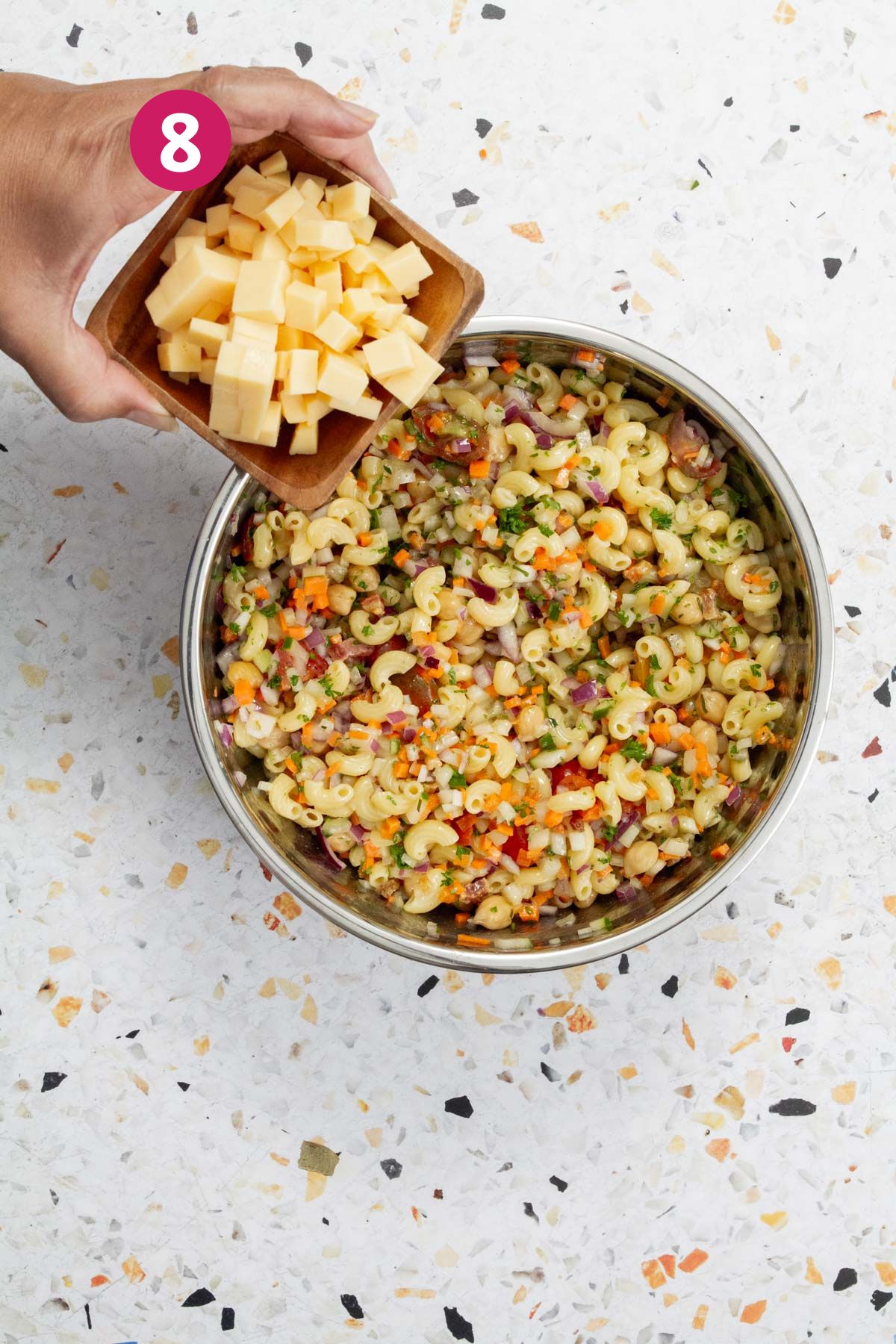 Hands adding cubed cheese from a wooden bowl into pasta salad with vegetables and chickpeas.