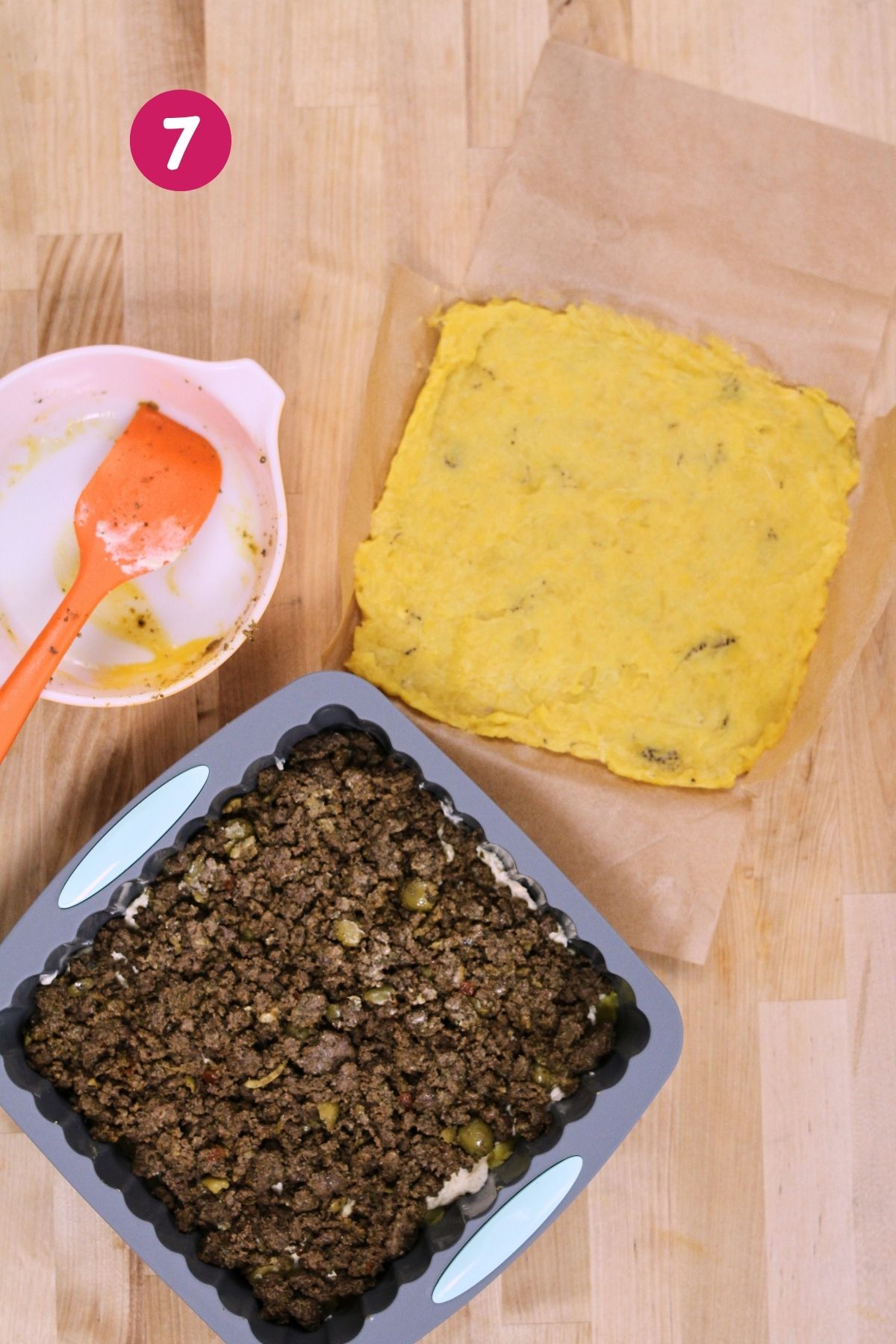 (Step 7) Seasoned picadillo spread in the baking pan next to the pre-shaped top mashed plantain layer on parchment paper, with the egg wash bowl and spatula visible.