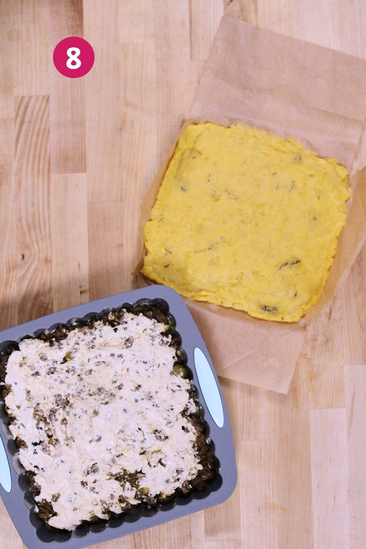 (Step 8) Shredded cheese spread over the picadillo layer in the baking pan, with the mashed plantain top layer waiting on parchment paper alongside.