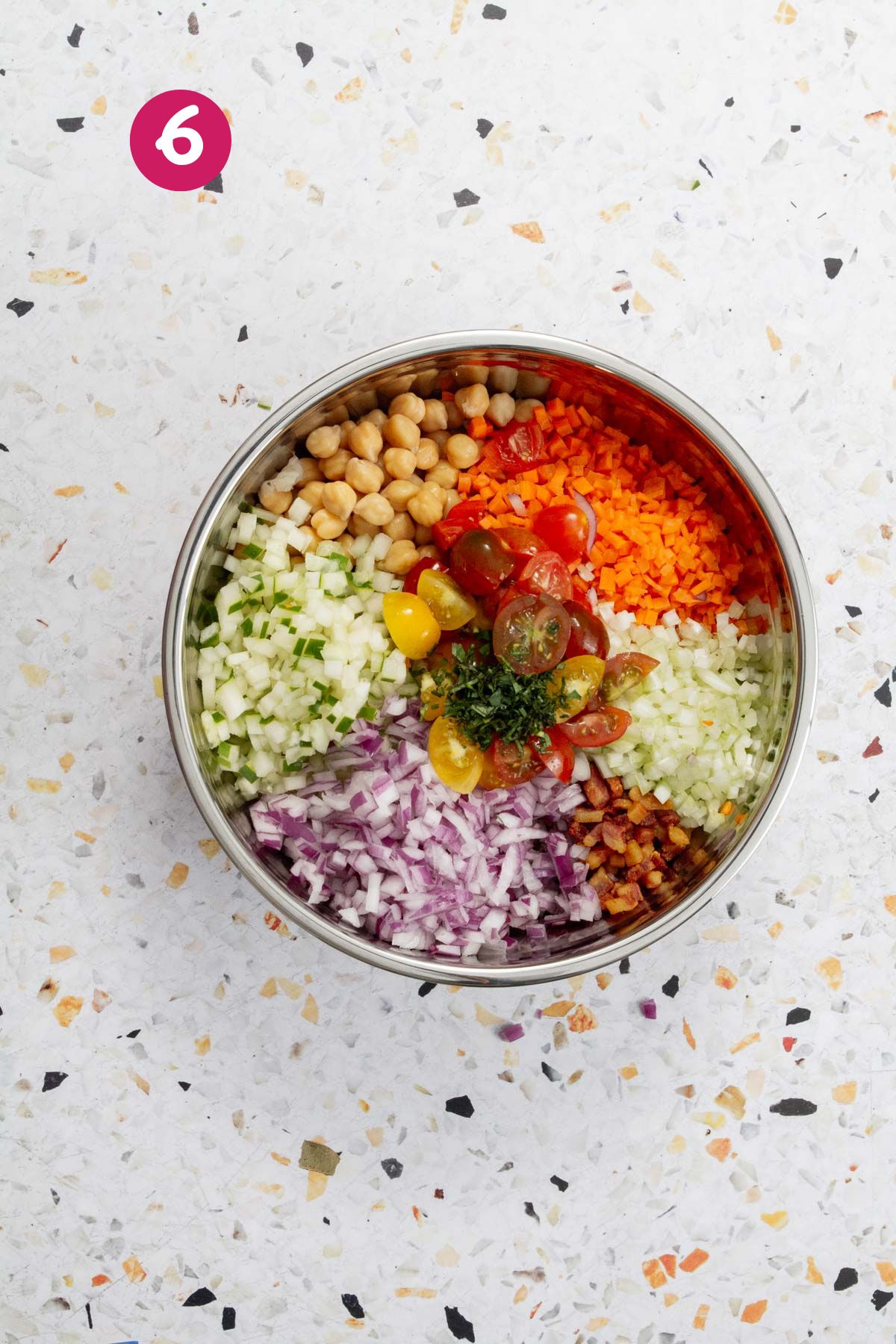 All pasta salad ingredients are arranged in a metal bowl, including chickpeas, diced vegetables, cherry tomatoes, celery, carrots, and red onion.