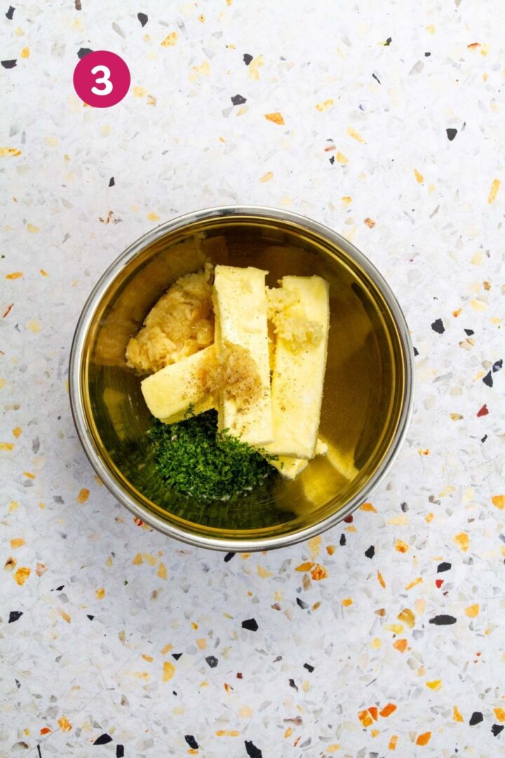 Butter sticks, roasted garlic paste, chopped parsley, and black pepper are combined in a stainless steel bowl before mixing.