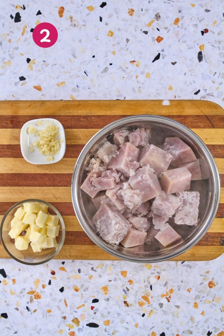 Cooked malanga cubes with minced garlic and cubed butter in a stainless steel mixing bowl, ready to mash.