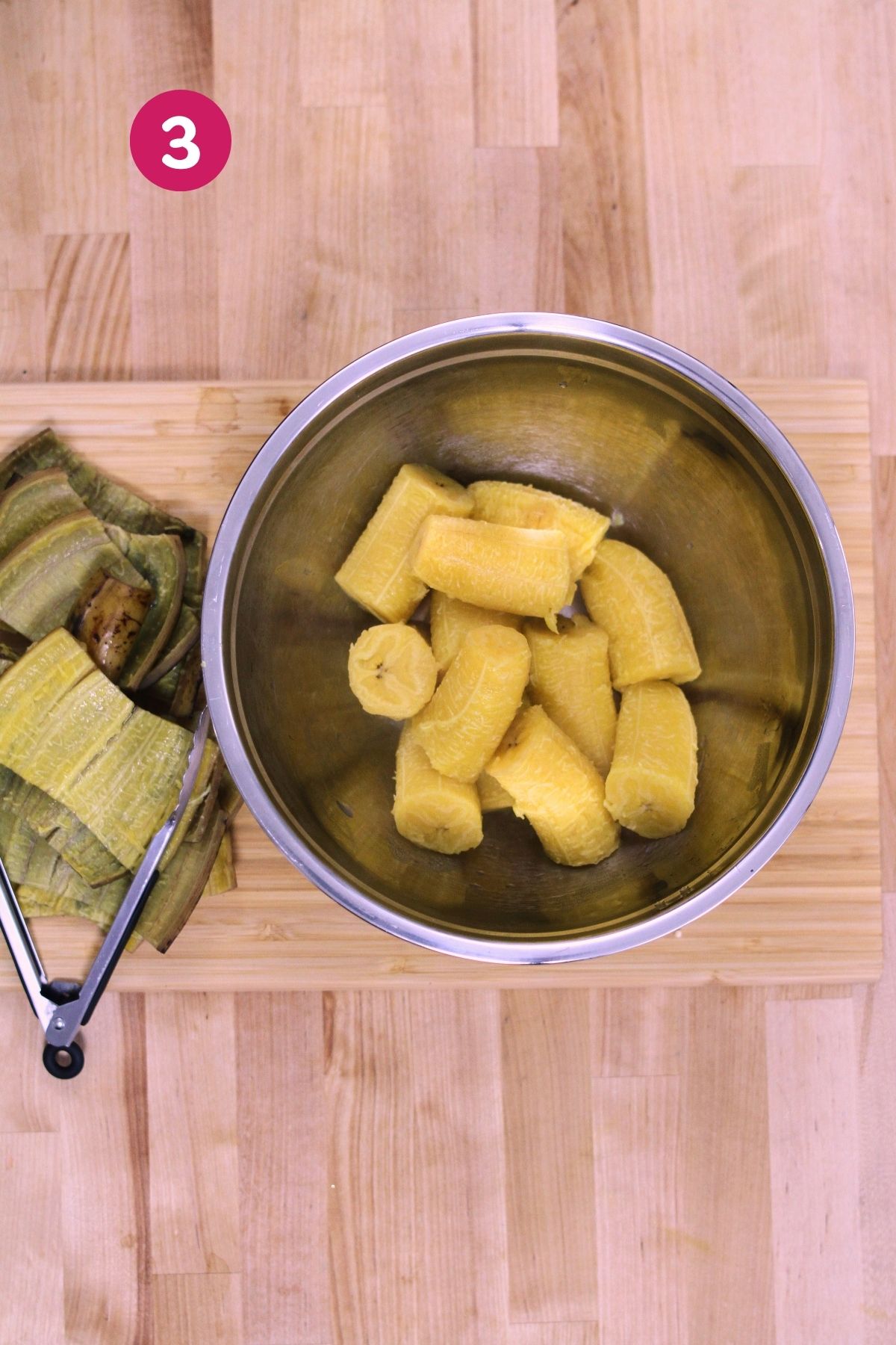 (Step 3) Peeled boiled plantain pieces in a stainless steel mixing bowl, ready to be mashed, with discarded skins on a cutting board alongside.