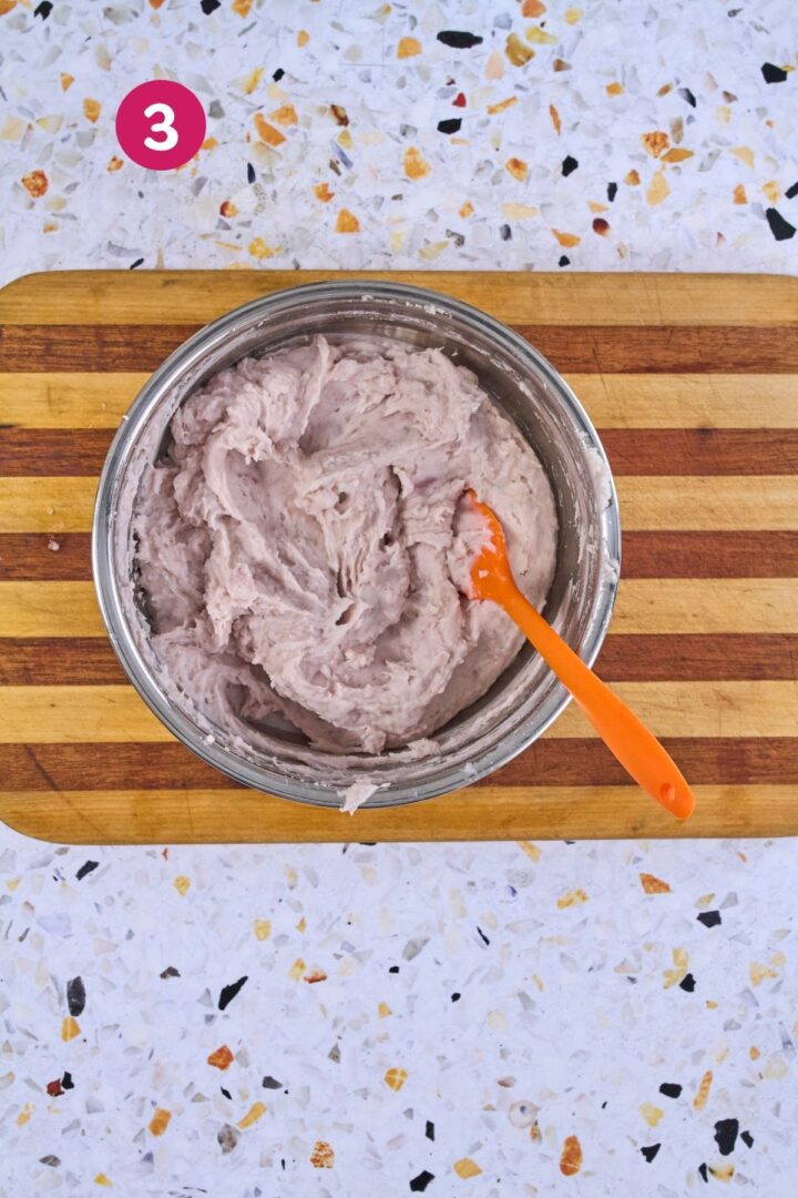 Finished mashed malanga in a stainless steel bowl with an orange spatula on a striped wood cutting board.