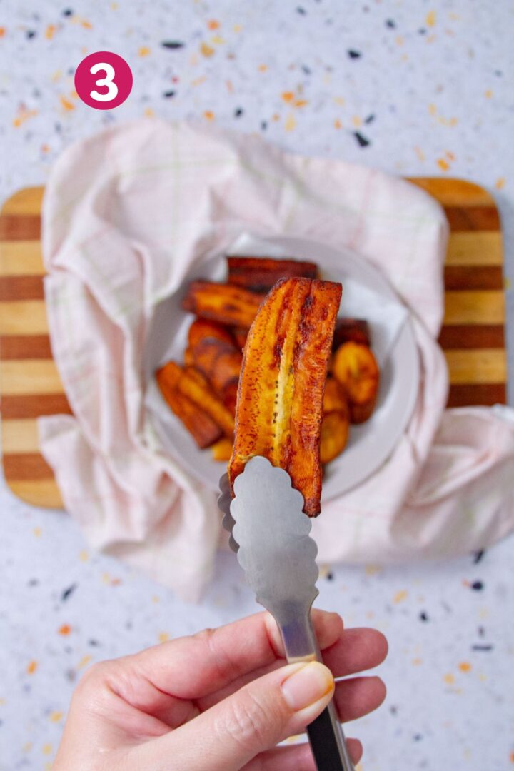 A long caramelized sweet plantain strip held upright in tongs, showing its deeply browned edges and soft interior, with a bowl of amarillos blurred in the background.