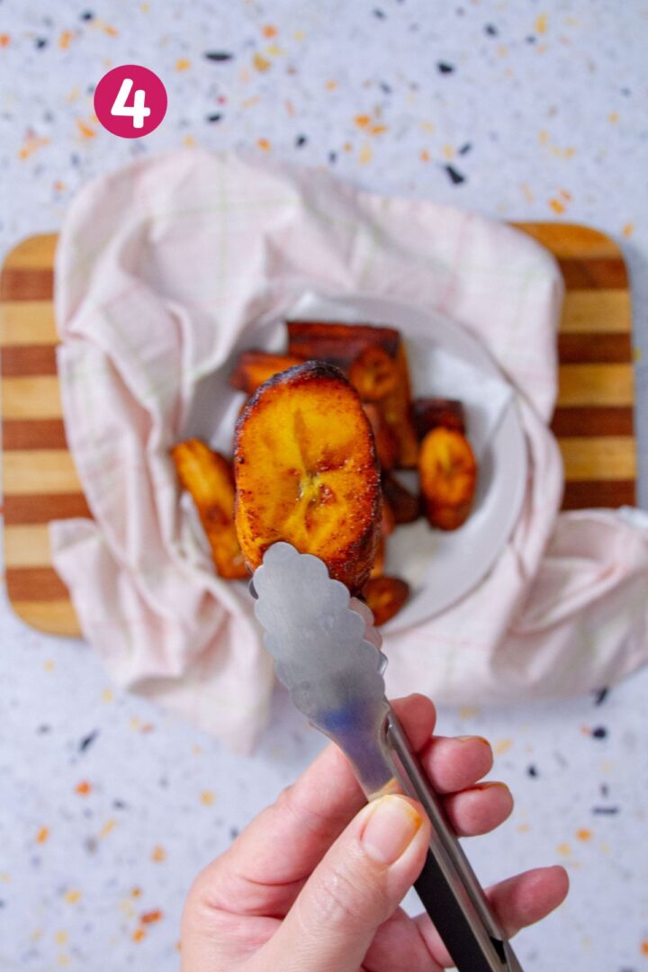 A round golden-brown fried sweet plantain slice held in tongs close to the camera, with a cloth-lined bowl of amarillos on a striped cutting board softly blurred in the background.