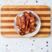A white bowl filled with caramelized fried sweet plantains rests on a striped wooden cutting board against a speckled terrazzo surface.